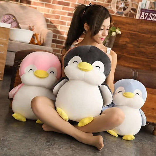 Three large plush penguin toys in pink, gray/white, and light blue are arranged on a floor in front of a seated woman in a cozy indoor room with brick and wood walls.