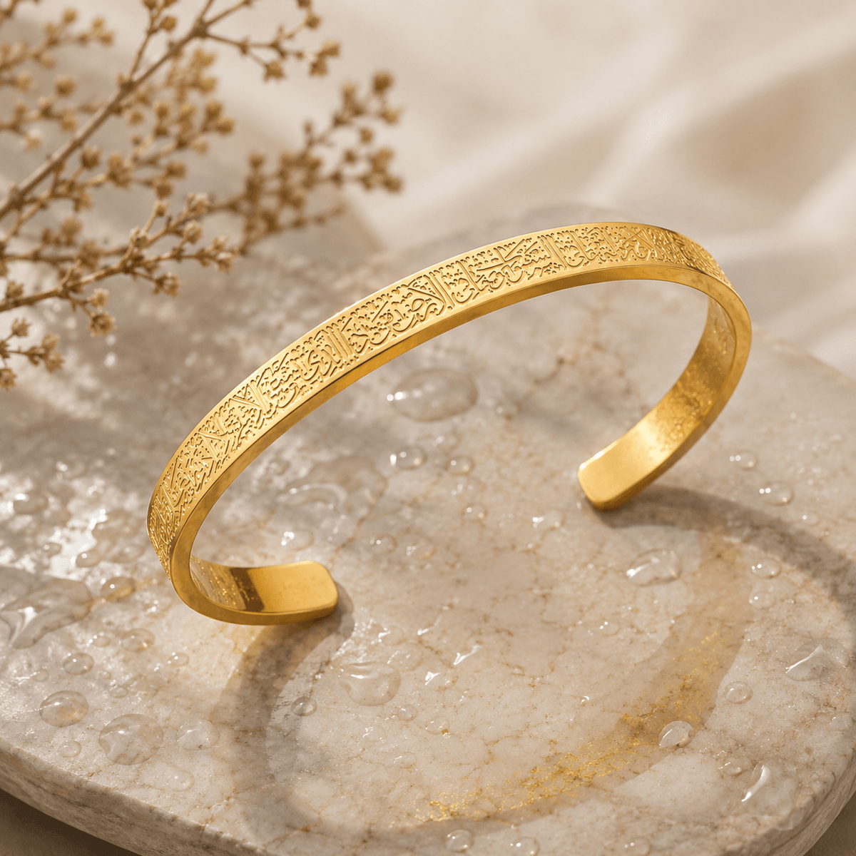 A gold-toned open cuff bracelet with intricate Arabic calligraphy engraving, displayed on a textured stone slab with dried flowers in the background.