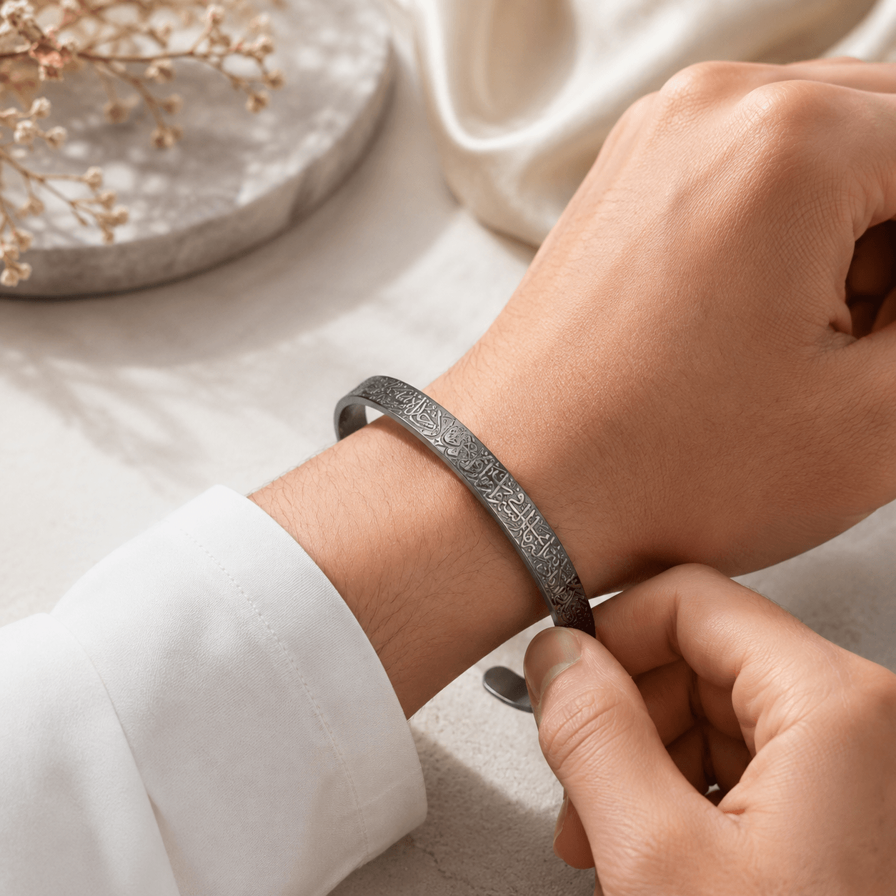 A close-up of a person's wrist wearing a textured, engraved silver-toned metal cuff bracelet, with another hand adjusting it. The background is a soft, light beige surface with a blurred decorative stone and white fabric.