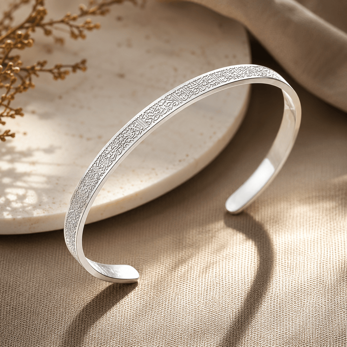 A polished silver-tone open cuff bracelet with an intricate engraved pattern along the outer curve, displayed on a textured beige fabric surface next to a light stone dish.