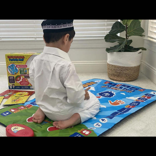 A young child wearing a white shirt and black skullcap sits on a colorful interactive floor mat in a bright room, with a toy box and books nearby and a potted plant in the background.Interactive Islamic prayer mat for kids